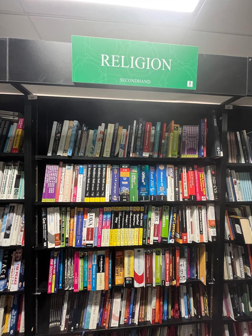 A bookcase in a second hand book store, labeled “religion”. On it, are old books about computer software and programming languages 