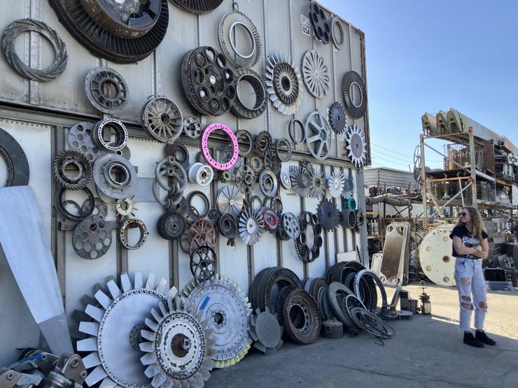 Robyn looking over her shoulder at a wall of turbine blades, things that look like hubs, basically beautiful metal round objects ranging from 6 inches to 5 feet in diameter, hung in random order with striking pink round somethings in the middle.